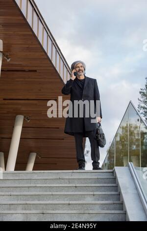 Confident bearded grey-haired businessman in coat using smartphone while consulting client or making appointment on top of staircase Stock Photo