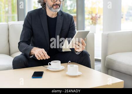 Mature bearded broker in formalwear having coffee and looking through online news while spending time in cafe of modern business center Stock Photo