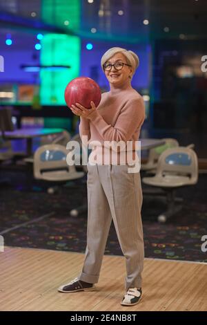 Vertical full length portrait of smiling senior woman holding bowling ball and looking at camera while enjoying active entertainment at bowling alley Stock Photo
