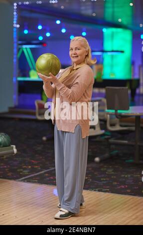 Vertical full length portrait of elegant senior woman holding bowling ball and smiling at camera while enjoying active entertainment at bowling alley Stock Photo