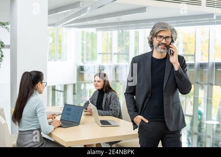 Confident mature businessman in formalwear talking to client on mobile phone against two female co-workers working over project by desk Stock Photo