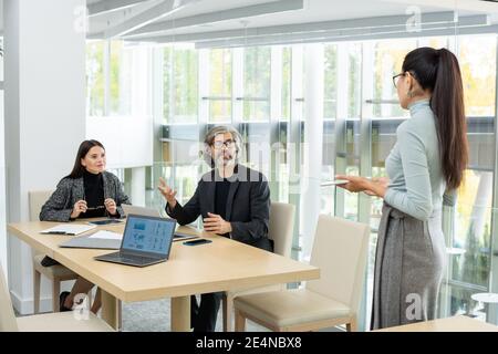 Two modern elegant economists looking at their Asian female colleague during discussion of graphic financial data at meeting in office Stock Photo