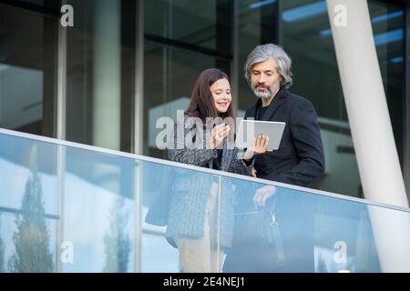 Young smiling female with touchpad showing and explaining online data to mature elegant businessman in black coat by office center Stock Photo