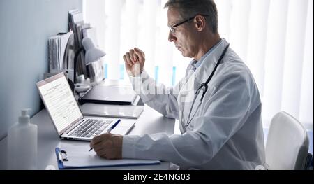 Serious older doctor using laptop computer in medical office. Stock Photo