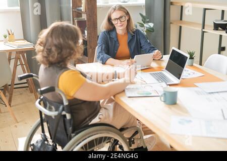 Two young contemporary managers sitting by table and looking at one another during discussion of financial data and planning work Stock Photo