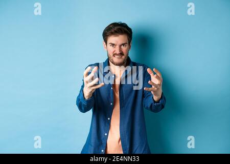 Angry tensed man wants to strangle someone, stretch out hands to choke enemy, biting lip and looking furious, standing on blue background Stock Photo