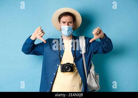 Covid-19 and summer holidays concept. Disappointed tourist in medicla mask frowning, showing thumbs down, negative feedback about travel, standing on Stock Photo