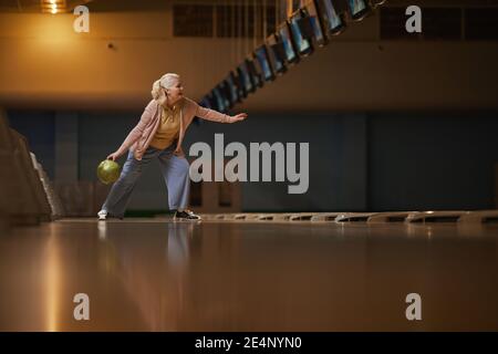 Wide angle side view at senior woman playing bowling alone while enjoying active entertainment at bowling alley, copy space Stock Photo