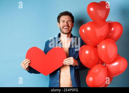 Heartbroken man crying of breakup of valentines day, holding red heart cutout and standing near romantic balloons over blue background Stock Photo