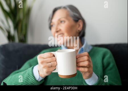 A close-up a mug in hands of a charming senior mature lady in out of focus, looking in the window, daydreaming and holding a hot drink, sitting on the