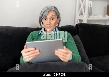 A surprised senior mature woman sitting on the couch, holding a pad and reading news online, looking at the tablet and cannot believe her eyes Stock Photo