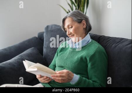 Charming grandmother senior mature woman in a green sweater, sitting on the couch and reading a book, middle-aged lady enjoying her leisure free time Stock Photo