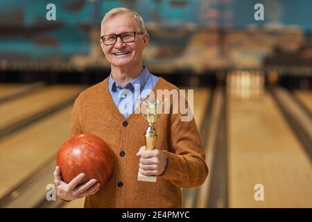 Waist up portrait of smiling senior man holding trophy and bowling ball while posing at bowling alley after winning match, copy space Stock Photo