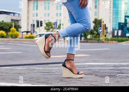 Woman in blue stylish denim jeans and wedge sandals with 1 leg raised in the city street. Stock Photo