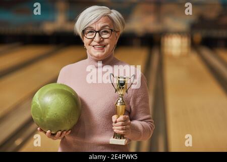 Waist up portrait of smiling senior woman holding trophy and looking at camera while standing in bowling alley after winning match, copy space Stock Photo