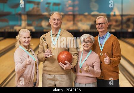 Waist up portrait of smiling senior team wearing medals holding trophy and looking at camera while standing in bowling alley after winning match Stock Photo