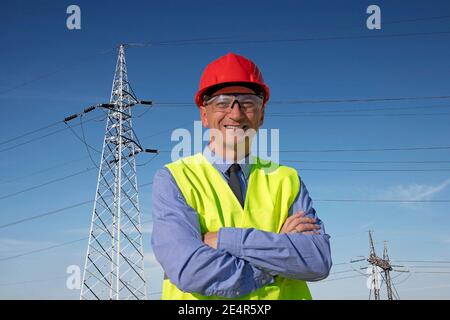 Smiling Engineer in Red Hardhat and Yellow West Standing Under The Electrical Transmission Towers. Happy Manager in Red Hardhat and Yellow Vest. Stock Photo
