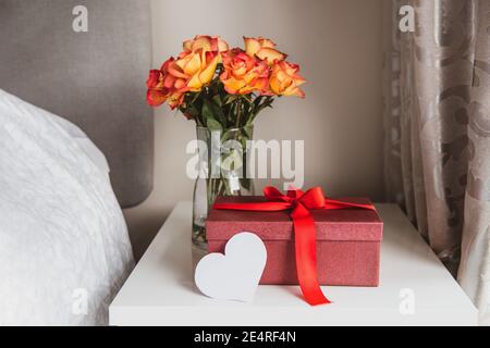 A beautiful gift box with red ribbon, a bouquet of roses flowers in vase and heart shaped blank greeting card on the bedside table against a gray wall Stock Photo