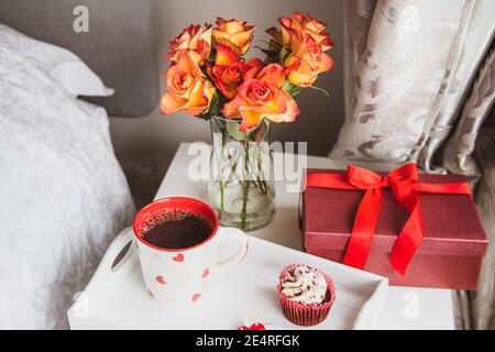 Tray with hot tea, cupcake, gift box and a bouquet of roses flowers in vase on the bedside table against a gray wall. Valentine's day, birthday, women Stock Photo