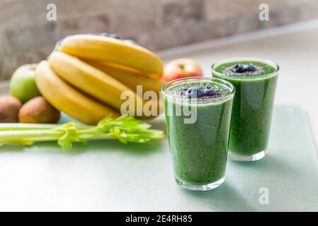 Two glasses of fresh green smoothie decorated with chia seeds and blueberry on the kitchen table with ingredients. Healthy food, detox eating Stock Photo