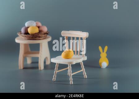 Creative Easter composition of yellow chocolate egg on a miniature wooden chair, easter bunny rabbit and nest with candy eggs on the table on the mute Stock Photo