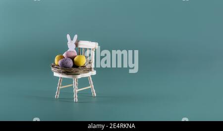 Creative Easter composition of small nest with candy chocolate eggs and Easter bunny rabbit on a miniature wooden chair on the muted dark green backgr Stock Photo