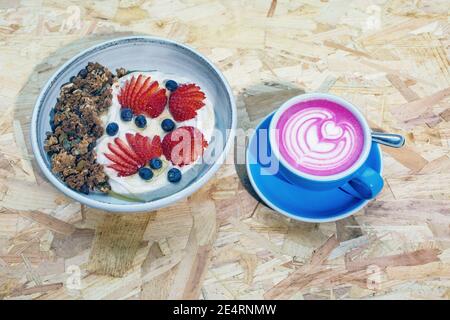 granola bowl with fresh fruits , yogurt and a pink rose beetroot latte in blue cup . Stock Photo