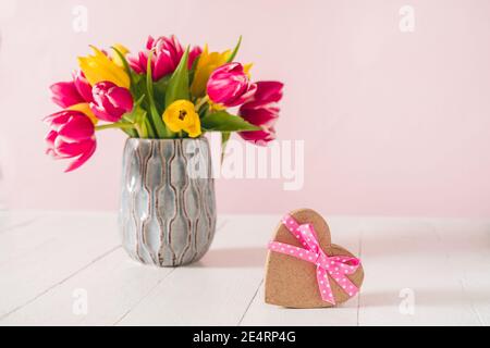 Craft heart shape gift box with satin ribbon bow and spring bouquet of pink and yellow tulip flowers in a vase on the white wooden table and pink Stock Photo