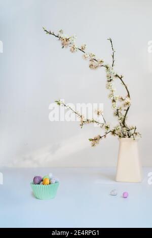 Blooming tree branch in a vase and small pastel colored Easter candy eggs in a paper bowl on light blue background. Soft selective focus. Happy Easter Stock Photo