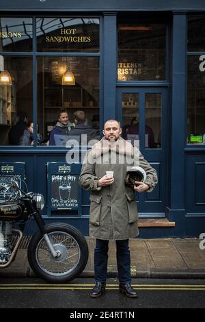 A man takes a coffee break at a cafe in London with classic motorcycle parked in front of a coffee shop Stock Photo