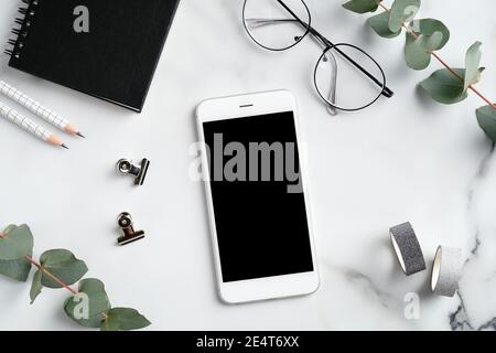 Modern feminine workspace with smartphone mockup, glasses, eucalyptus branches, notebook and office supplies on marble desk. Flat lay, top view. Elega Stock Photo