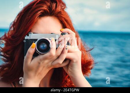 Red haired woman travel photographer, taking pictures on the background of the sea. Rare photographic camera for vacation snapshots. Closeup headshot Stock Photo