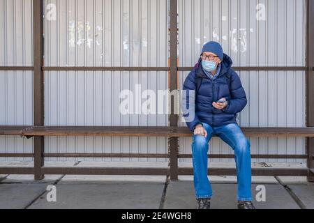 A middle-aged man in a medical mask sits at a public transport stop in the fall and waits for a bus. Using a smartphone, tracks location infirmation. Stock Photo