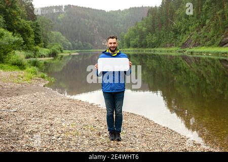Your text here. Handsome young man holding empty blank board outdoors with beautiful nature on background. Stock Photo