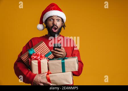 Excited young african american man in santa claus holding stack of boxes isolated on yellow background, using mobile phone Stock Photo