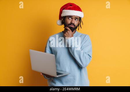 Pensive young santa african american man in Christmas hat working on laptop pc computer isolated over yellow background Stock Photo