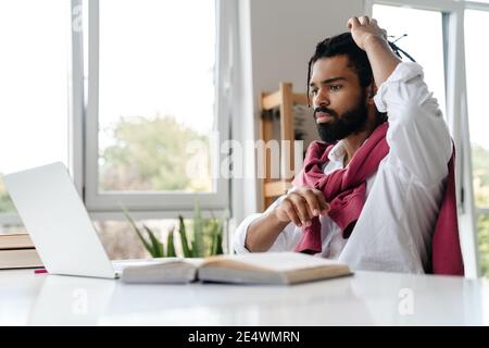Focused african american guy working with laptop and book while sitting in office Stock Photo