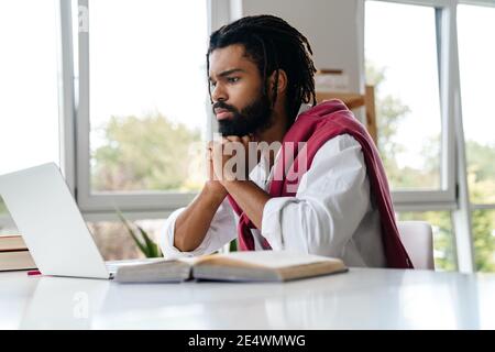Focused african american guy working with laptop and book while sitting in office Stock Photo