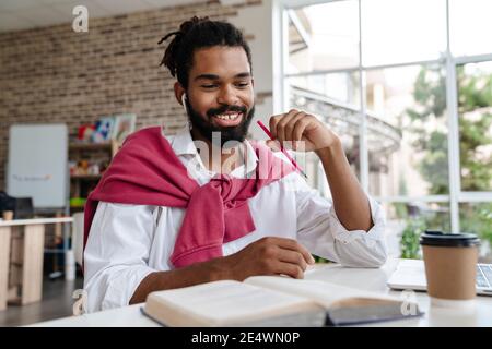 Joyful african american guy in earphones working with book and laptop while sitting in office Stock Photo