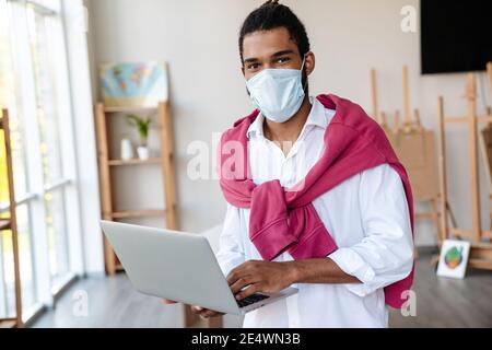 Joyful african american guy in face mask using laptop while standing at office Stock Photo