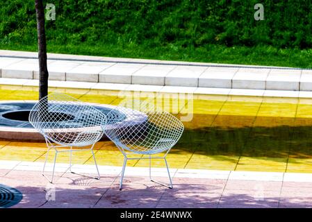 Two wicker chairs made of wire stand near a tree on a tile. Nobody is there, unusual chairs are standing in a park. Stock Photo