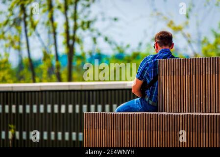 A young and beautiful millennial man sits on a bench in modern park at city. Back view. Stock Photo