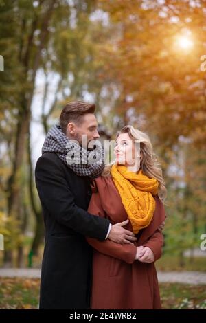 Ray of sun shining on the loving couple man and a woman hugged from behind smile looking at each other in the autumn park. Outdoor shot of a young Stock Photo