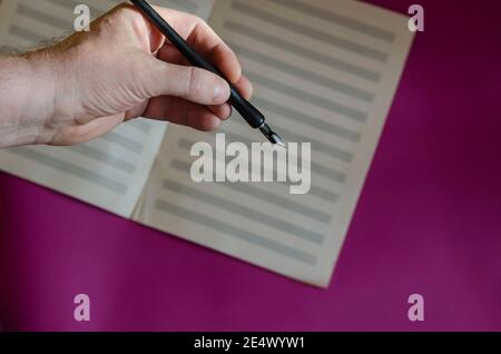 A man's hand with a black fountain pen and opening music notebook on a purple background. Musical creativity. Selective focus. Stock Photo