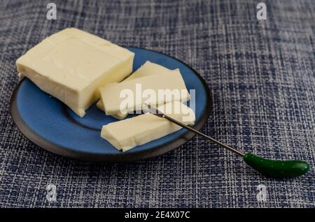 Rectangular slice of processed cheese in a blue saucer on a denim background. Sliced into rectangular portions. Skewer for stabbing. Dairy products, f Stock Photo