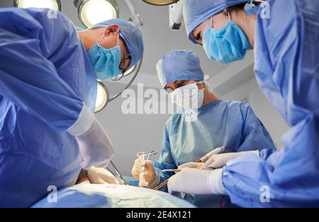 Surgical team in blue medical suits using medical instruments and performing plastic surgery. Male surgeon and female assistants wearing protective face masks, sterile gloves and caps in modern clinic Stock Photo