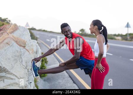 Fit african american man in sportswear stretching while woman is standing on a coastal road Stock Photo