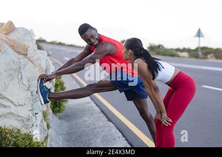 Fit african american man in sportswear stretching while woman is standing on a coastal road Stock Photo