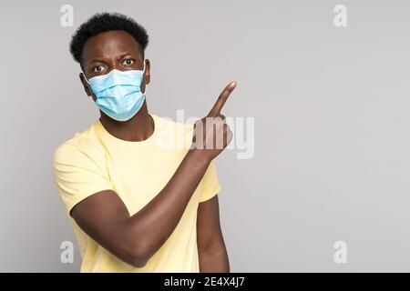 Afro man wear face mask points away with finger, showing at blank copy space. Covid-19 pandemic.  Stock Photo