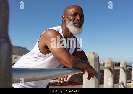Fit senior african american man leaning on fence against blue sky. healthy retirement technology communication outdoor fitness lifestyle. Stock Photo